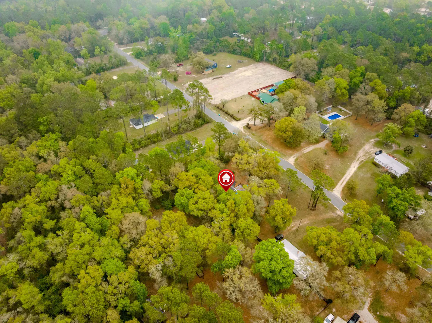 Aerial view of a wooded property with a small house marked by a red pin, surrounded by green trees and other houses in the distance.