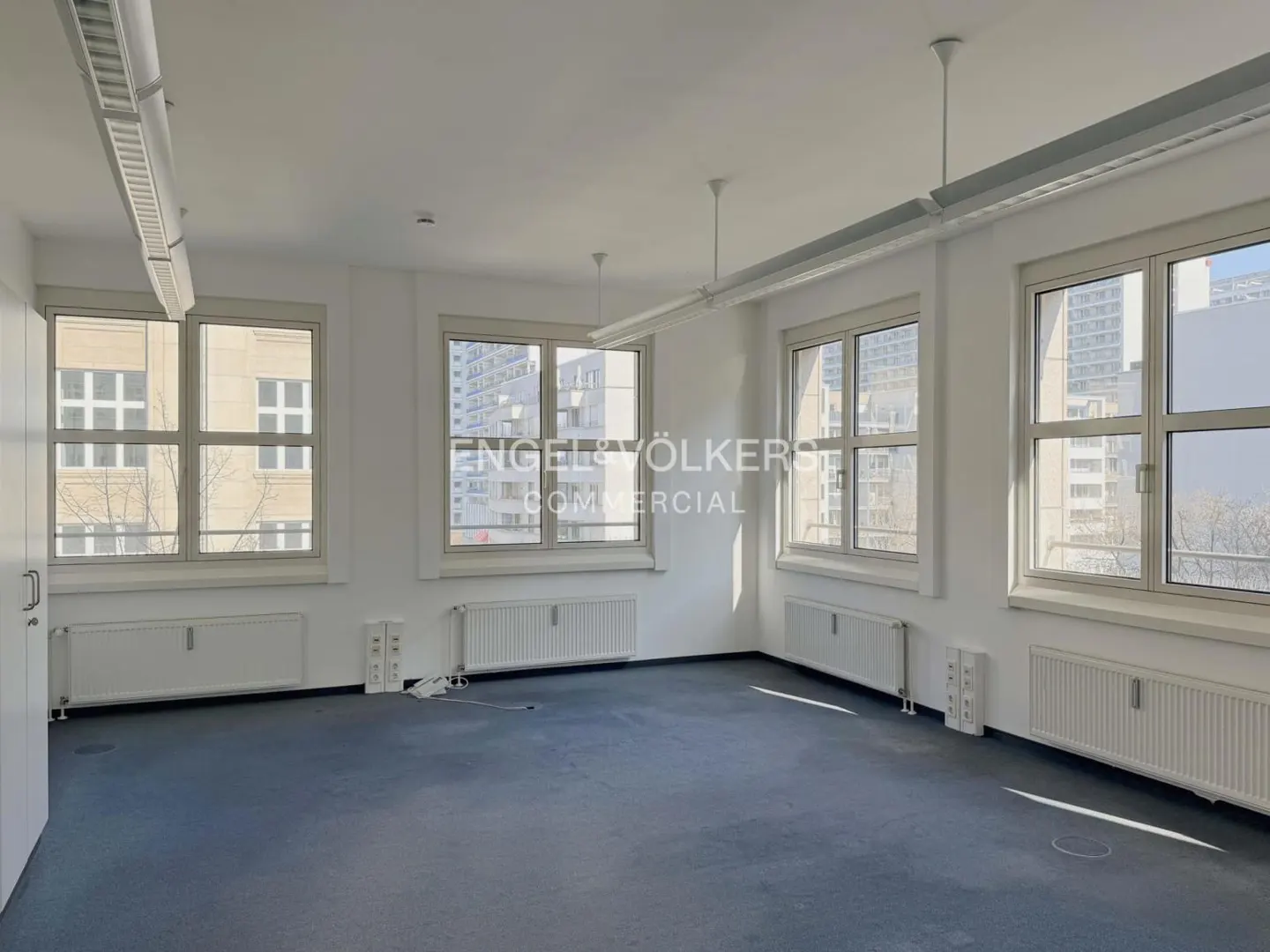 Bright, empty office space with blue carpet, white walls, and large windows overlooking city buildings. Radiators under windows.