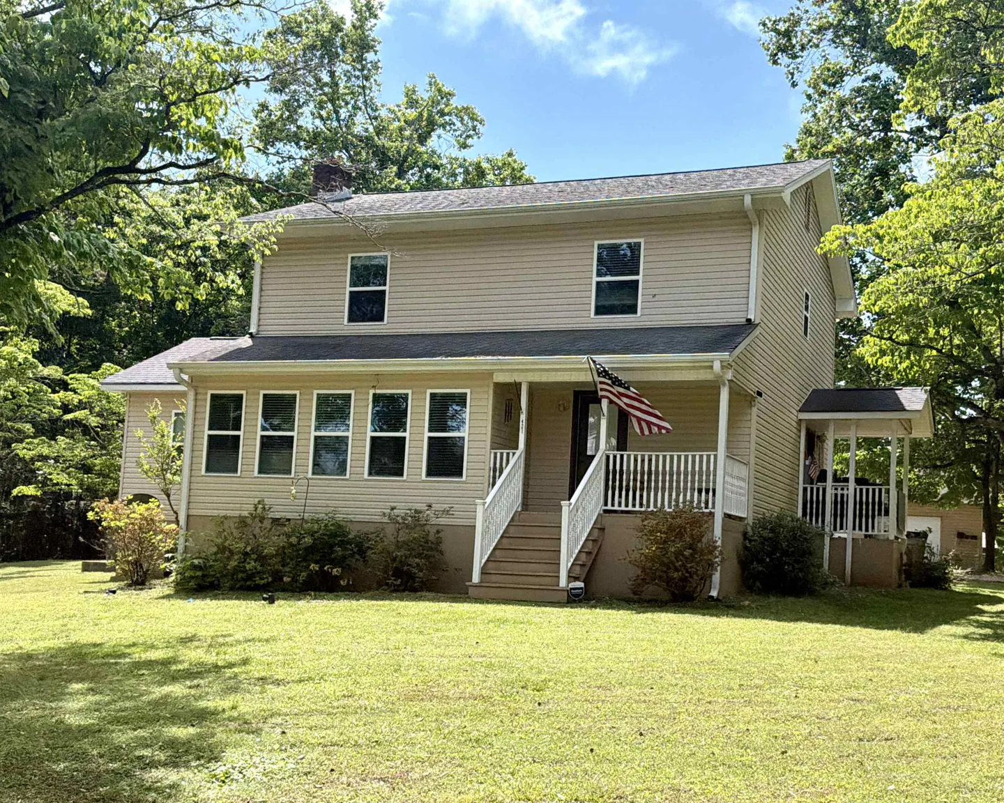 Two-story beige house with a front porch, white railings, and an American flag. Green lawn and trees surround the house.