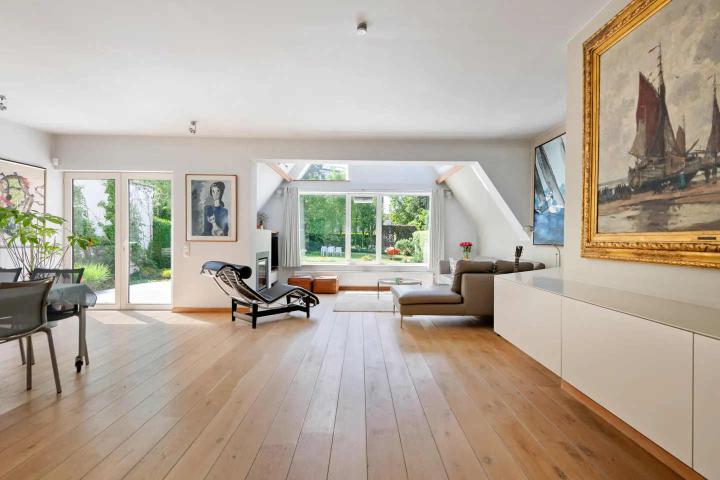 Bright, modern living room with wood floors, white walls, and large windows overlooking a green garden. Art adorns the walls, and a chaise lounge sits near a fireplace.