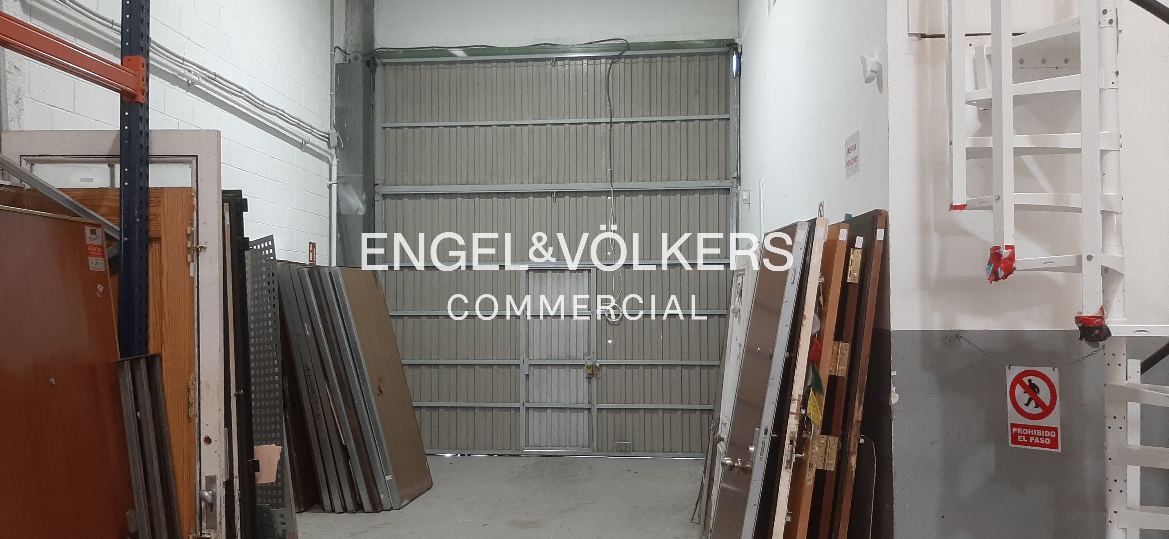 Interior of a warehouse with a large metal rolling door, stacks of doors leaning against walls, and a no-entry sign. [AI-generated text]