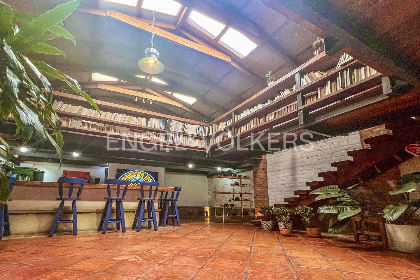 Rustic room with terracotta tile floors, a wooden bar with blue stools, a loft library, and a staircase under a skylight.