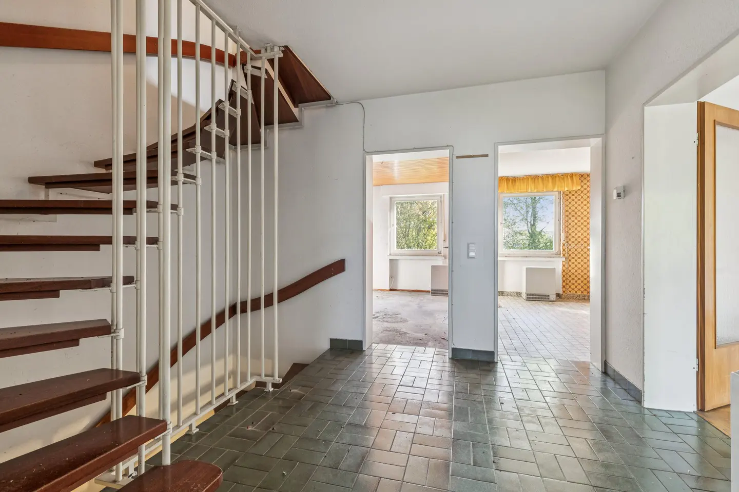 Interior view of a home's entryway with a brown wood staircase and white railing, leading to two open doorways. The floor is tiled in green.