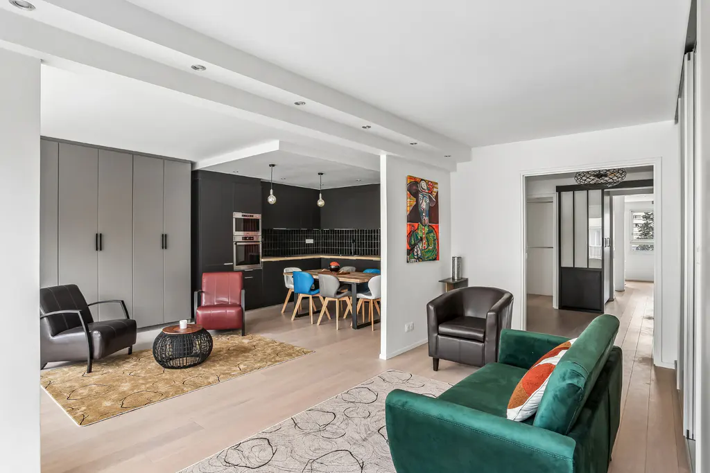 A modern apartment interior with a green sofa, chairs, and an open kitchen. The walls are white, and the kitchen has black cabinets and a dining table.