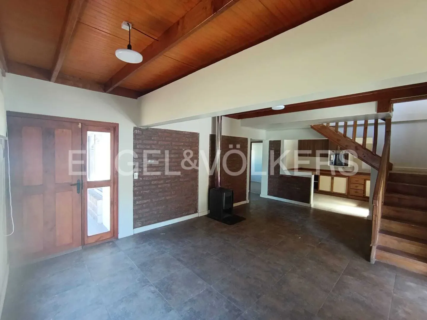 Open-plan living area with wood ceiling, brick accent wall, wood-burning stove, and staircase to the right.