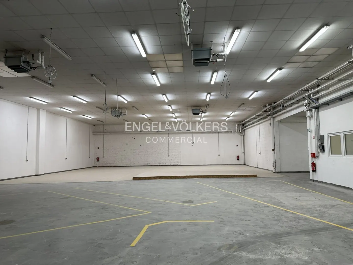 Spacious, empty commercial warehouse with white walls, concrete floors with yellow markings, and overhead fluorescent lights.