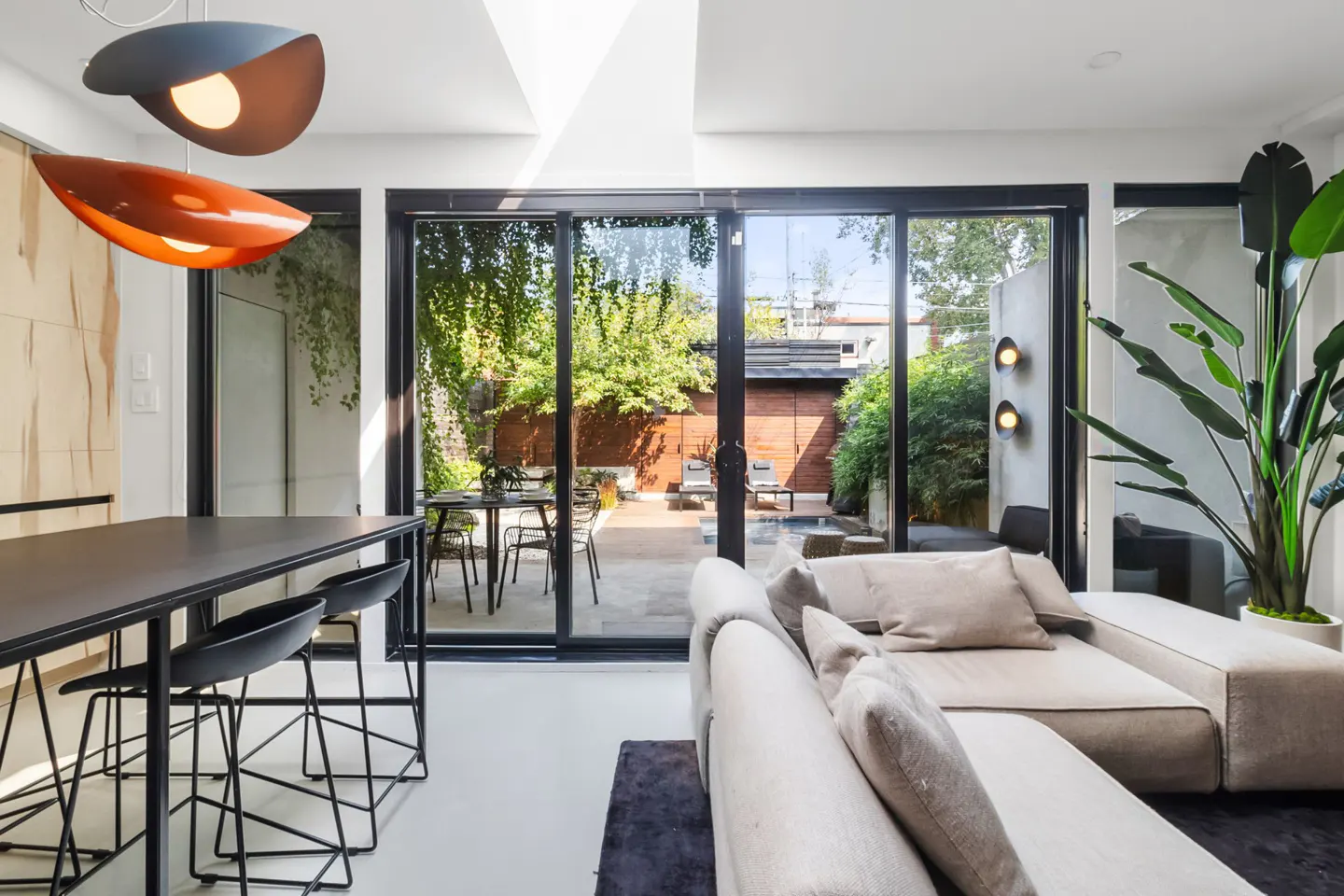 Bright living room with a black table, chairs, and a beige sectional sofa. Sliding glass doors open to a patio with greenery.