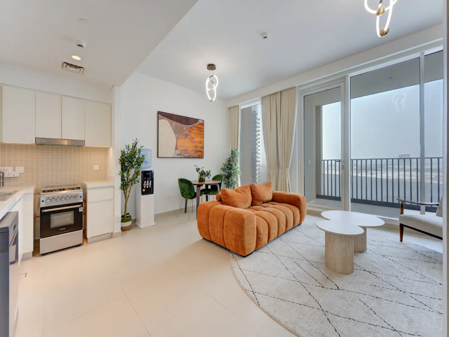 Bright apartment interior with an orange sofa, white rug, and balcony view. Kitchen area with white cabinets and stainless steel appliances.