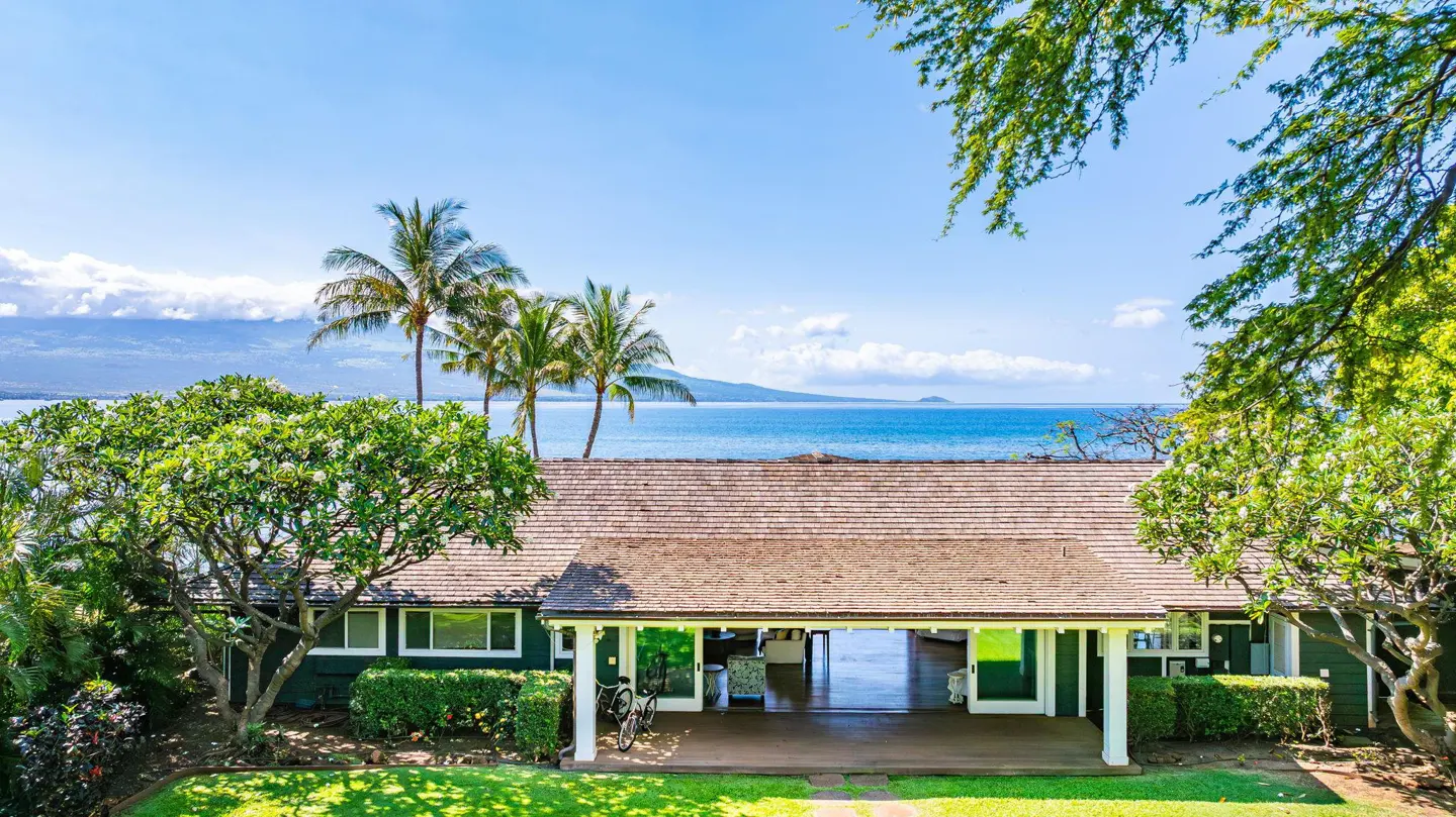 A tropical home with a shingled roof, open patio, and lush greenery, overlooking a blue ocean and distant mountains.