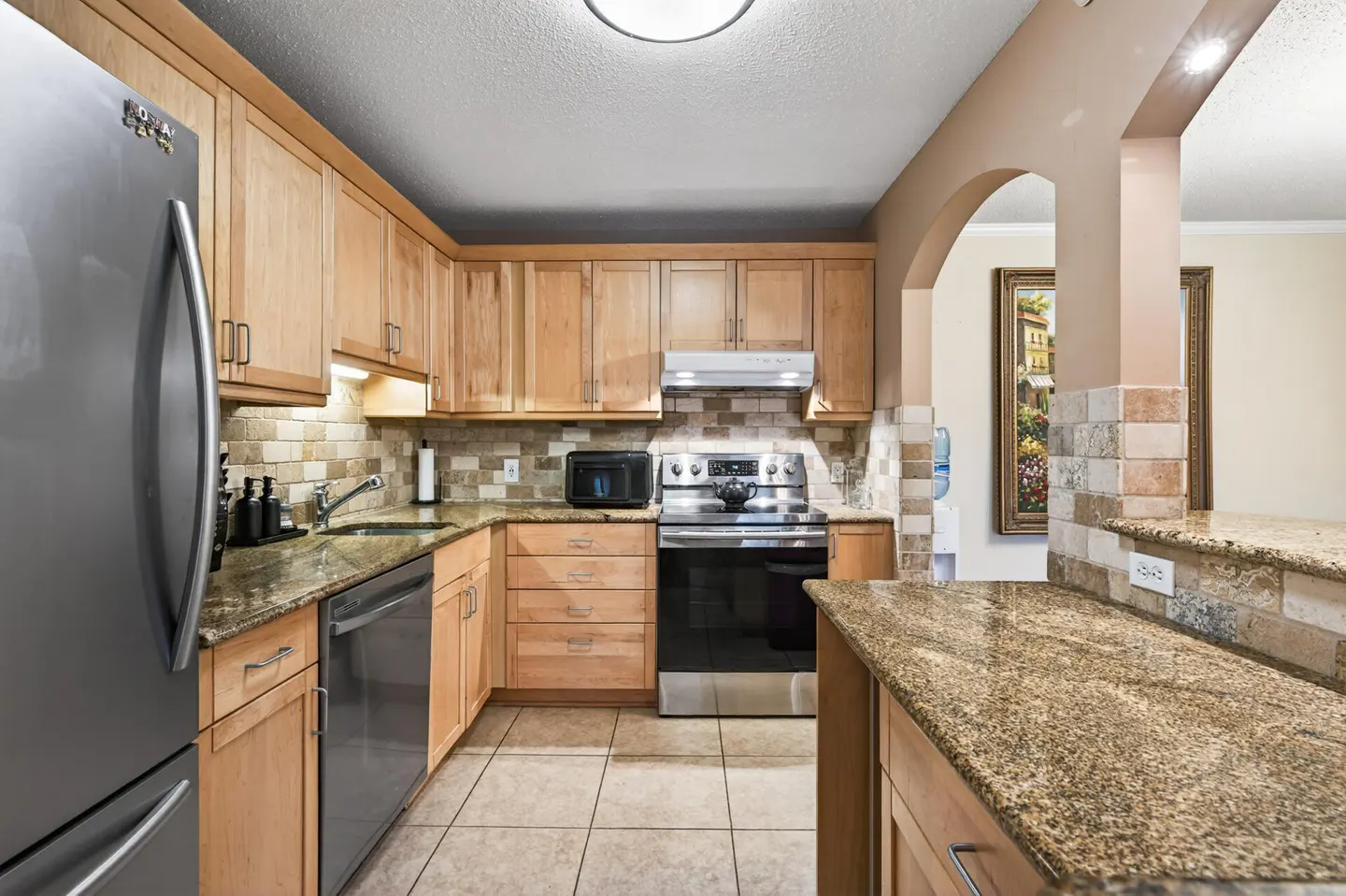 A kitchen with light wood cabinets, granite countertops, stainless steel appliances, and a tiled backsplash.