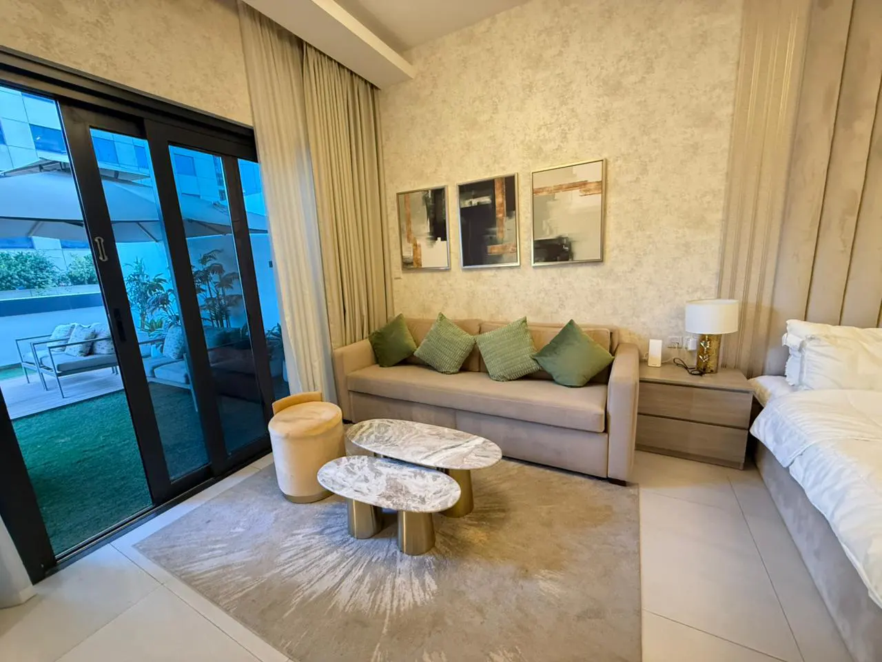 A modern living room with a beige sofa, green pillows, and marble coffee tables. Sliding glass doors open to a patio with outdoor seating.