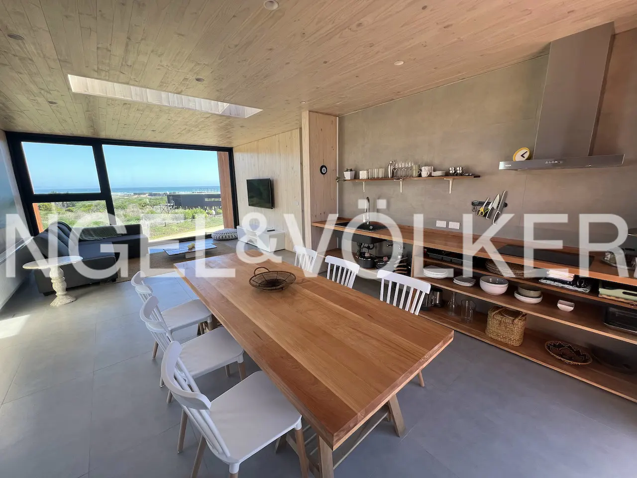 A modern dining area with a long wooden table and white chairs, a large window with an ocean view, and open shelving in the kitchen.