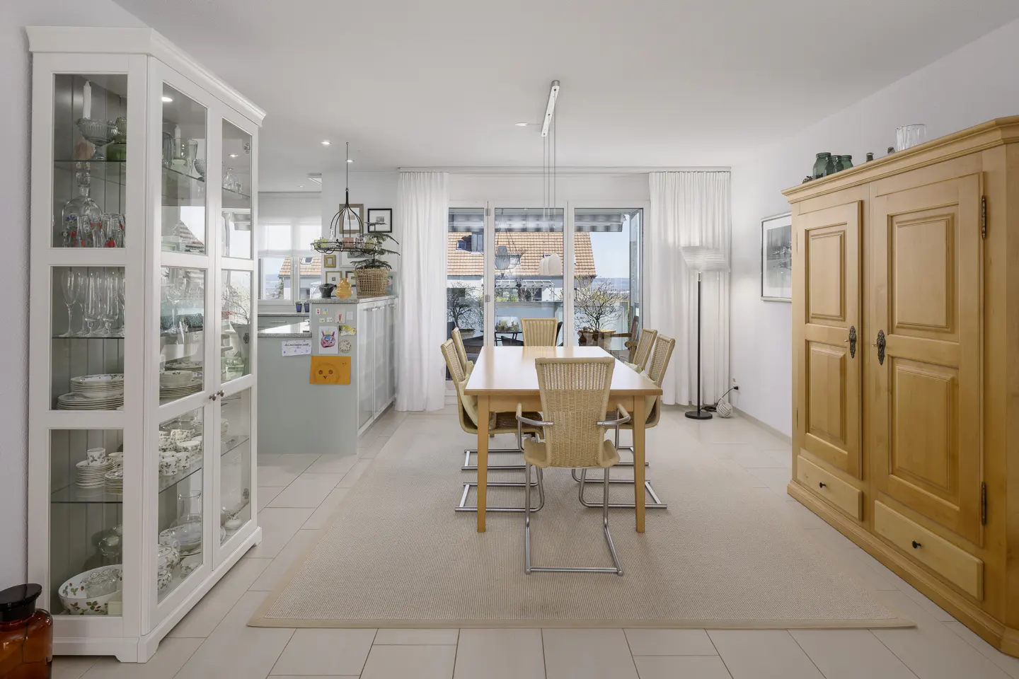 Bright dining room with a wooden table, six chairs, and a large wooden cabinet. A white china cabinet is on the left. Sliding glass doors lead to a balcony.