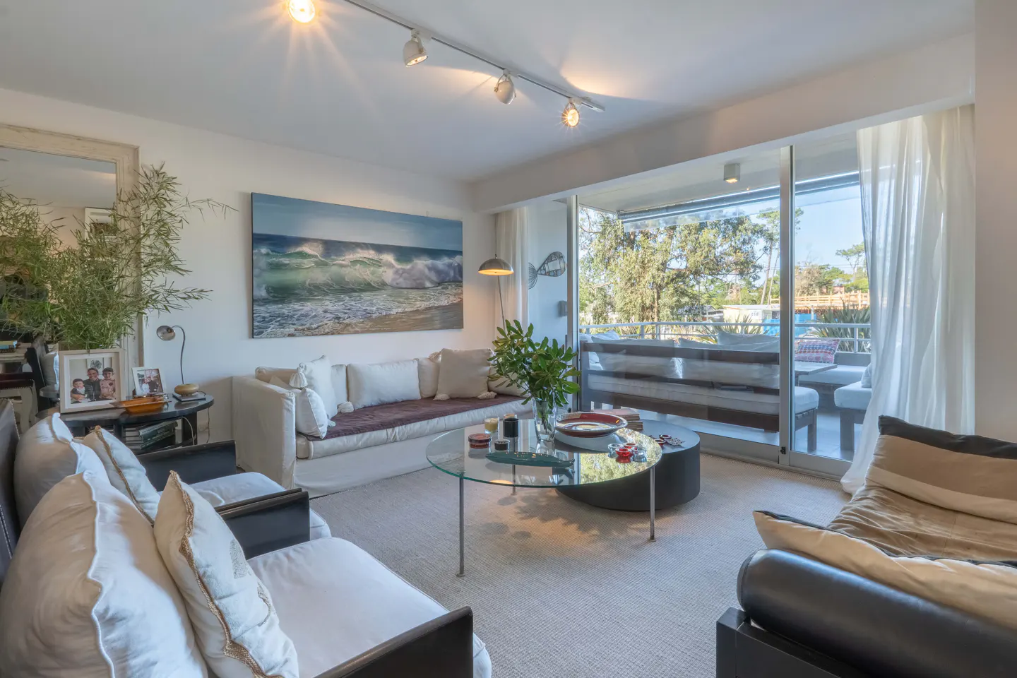 Bright living room with white sofa, chairs, and a glass coffee table. A large ocean wave painting hangs above the sofa. Balcony with outdoor seating visible.