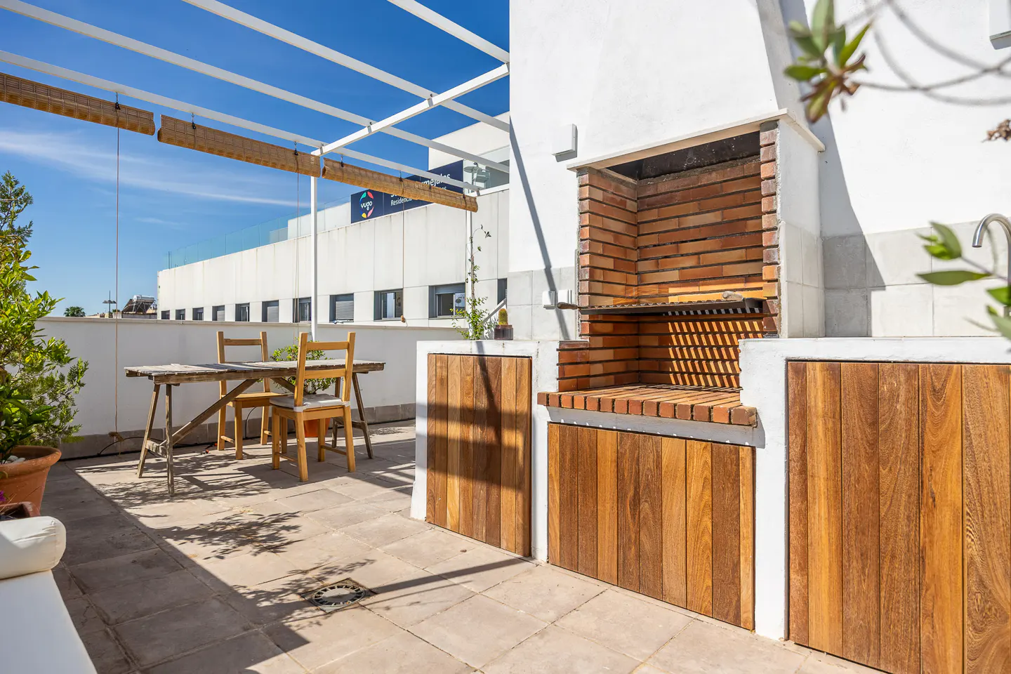 Rooftop patio with a brick barbecue, wooden cabinets, and a table with chairs under a white pergola.