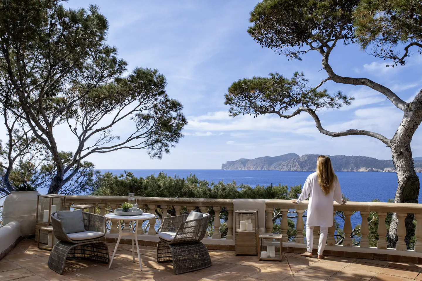 Woman on a terrace overlooking the ocean. Wicker chairs, table, and lanterns decorate the space. Trees frame the blue sky and water.