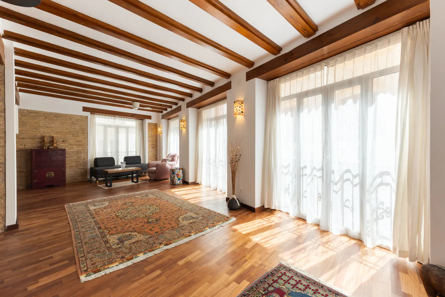 Bright living room with wood floors, exposed beams, and large windows with sheer white curtains. A patterned rug sits in the center.