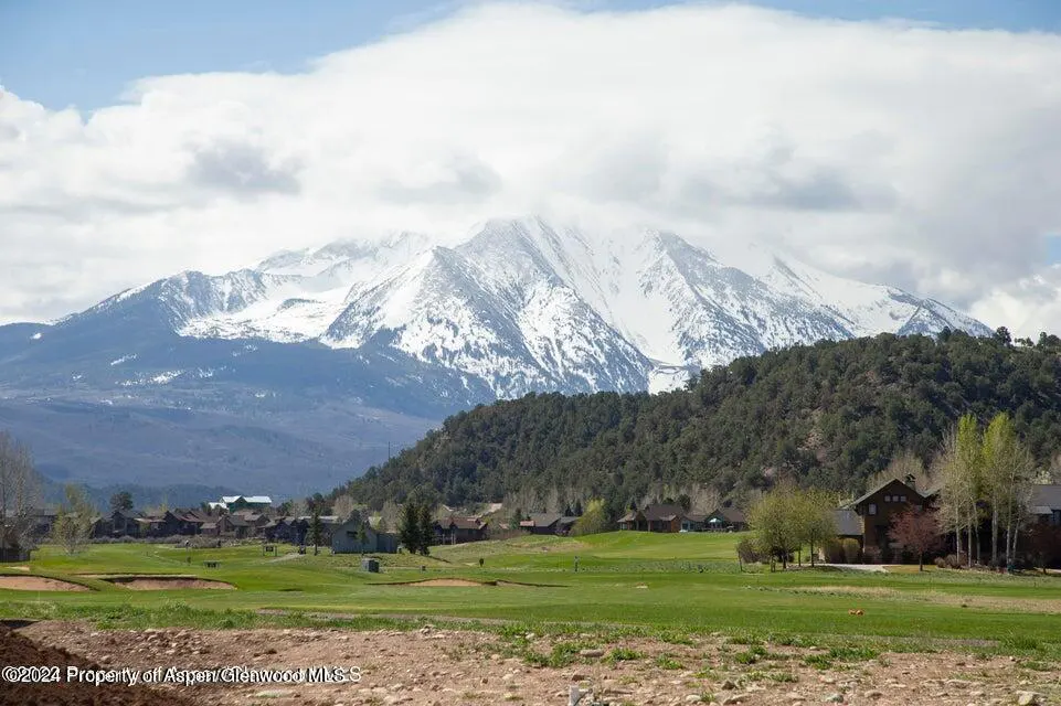 Scenic view of a green golf course with houses, trees, and a snow-capped mountain under a cloudy sky.
