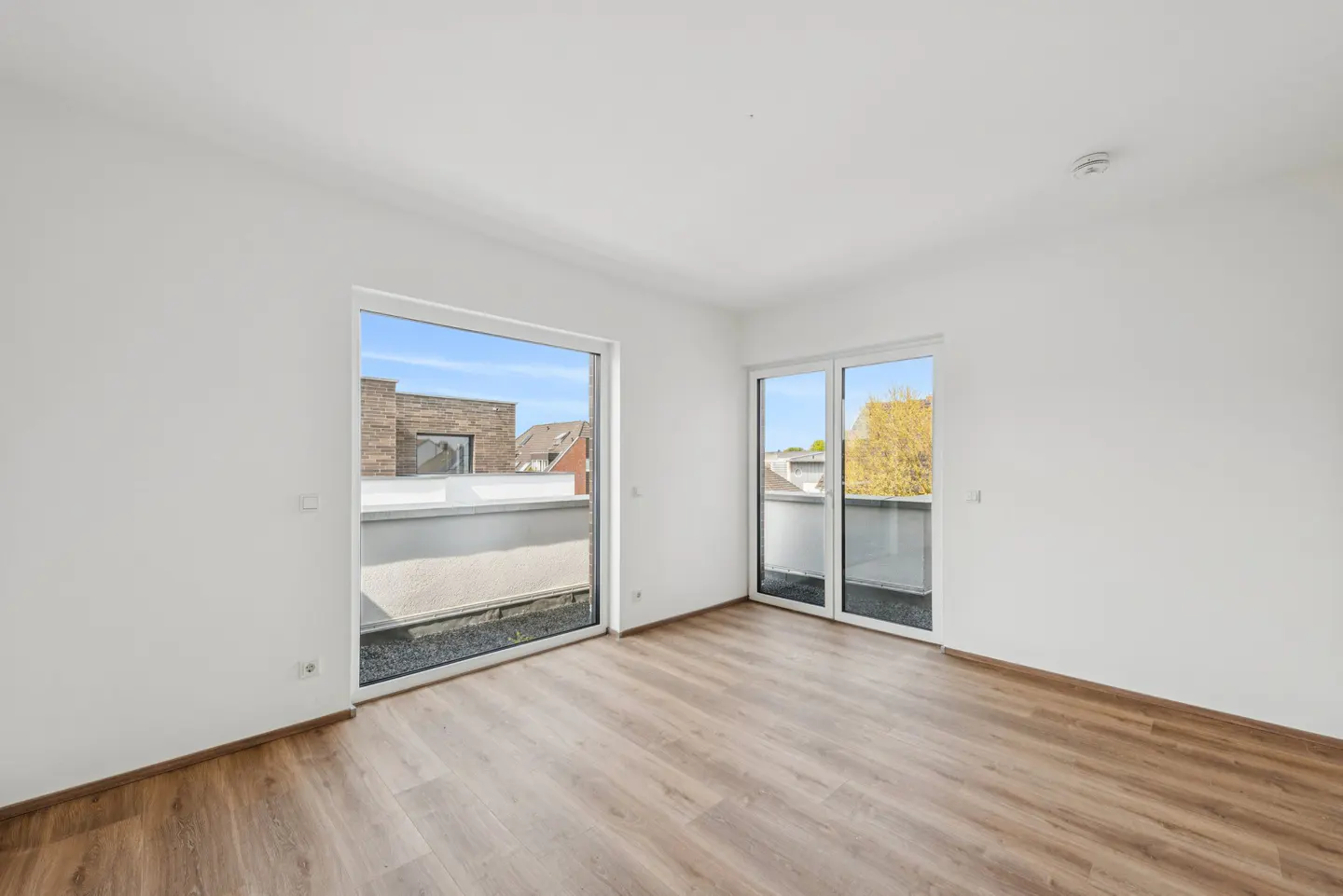 Bright, empty room with wood floors and white walls. Two glass doors open to balconies with views of neighboring buildings.