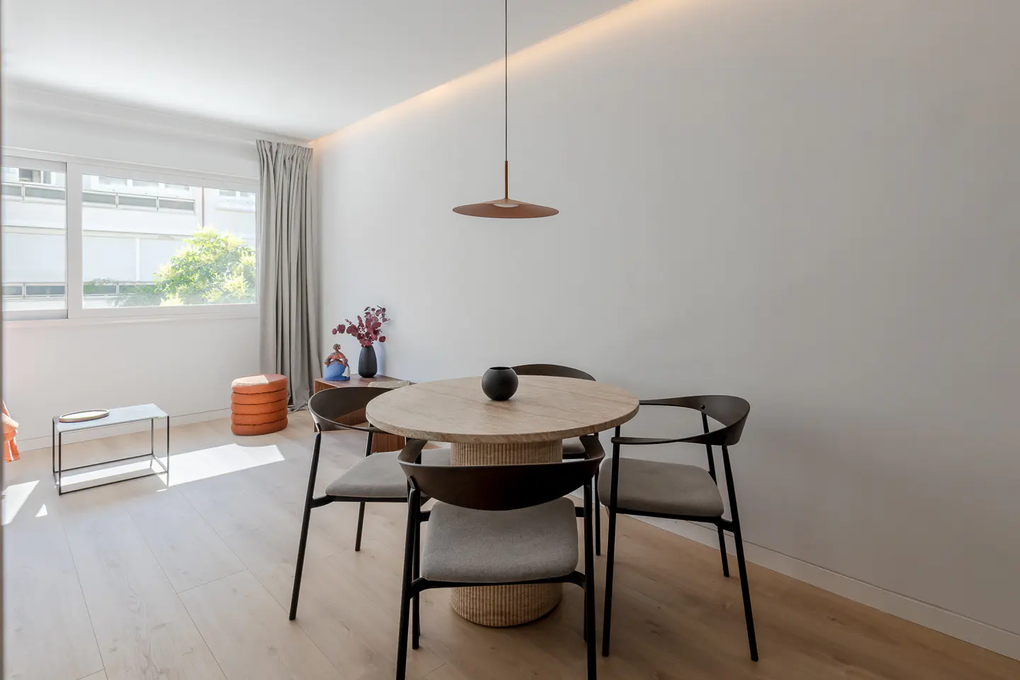 Bright dining area with a round table, four chairs, and a pendant light. A window with curtains is on the left.