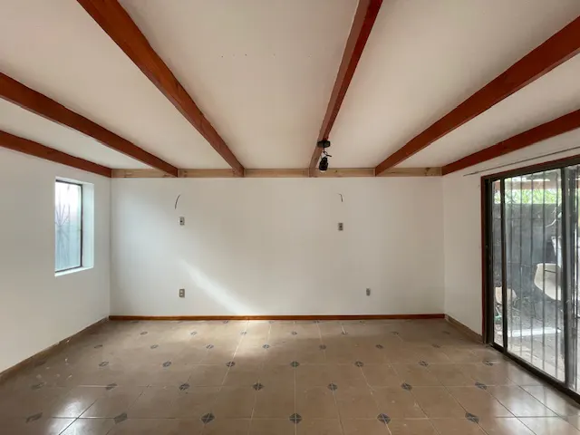 Empty room with white walls, tile floor, and exposed wood beams on the ceiling. A window and sliding glass door provide natural light.