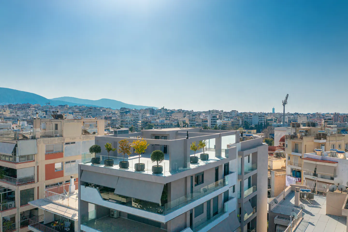 Aerial view of a modern grey apartment building with a rooftop terrace featuring potted trees, overlooking a city skyline.
