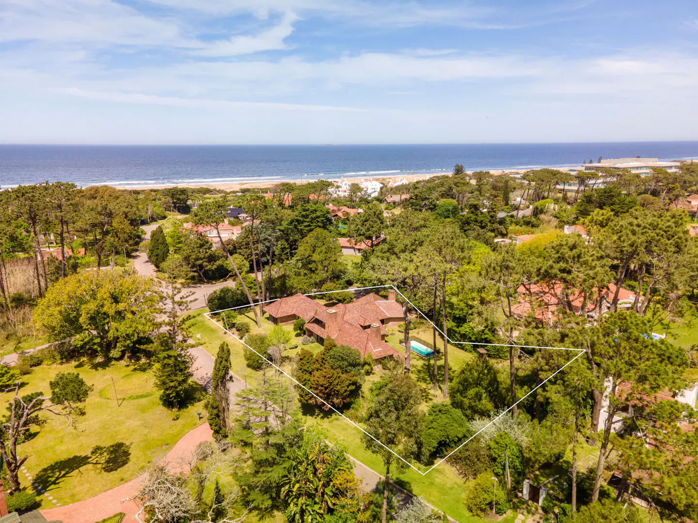 Aerial view of a property with a red-tiled roof, surrounded by green trees, with the ocean and beach in the background.