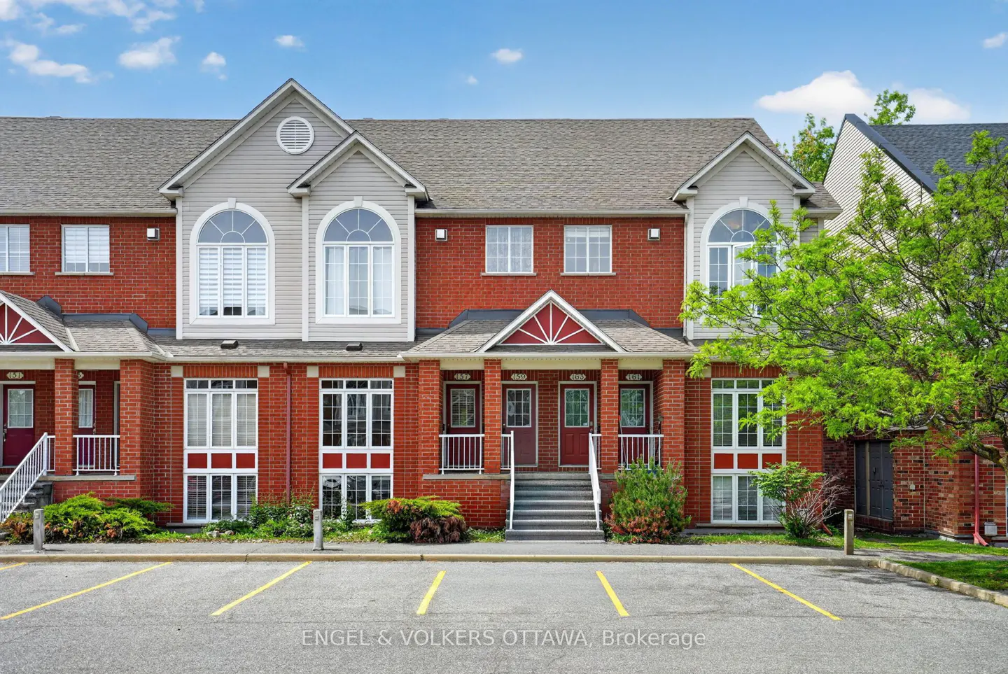 Exterior view of a red brick townhouse complex with grey siding, arched windows, and a paved parking lot in the foreground.