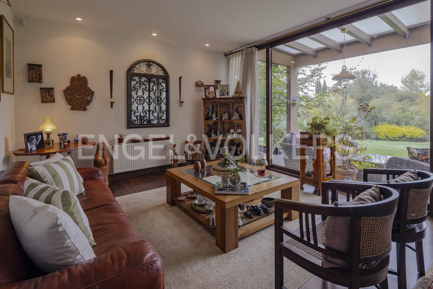 Living room with a brown leather sofa, wooden chairs, and a view of a green garden through a glass wall.