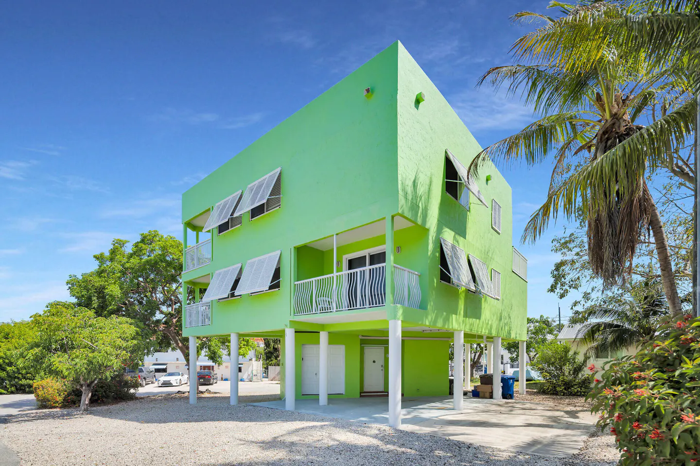 Two-story, bright green stilt house with white trim, balconies, and awning-style window covers under a blue sky with palm trees.