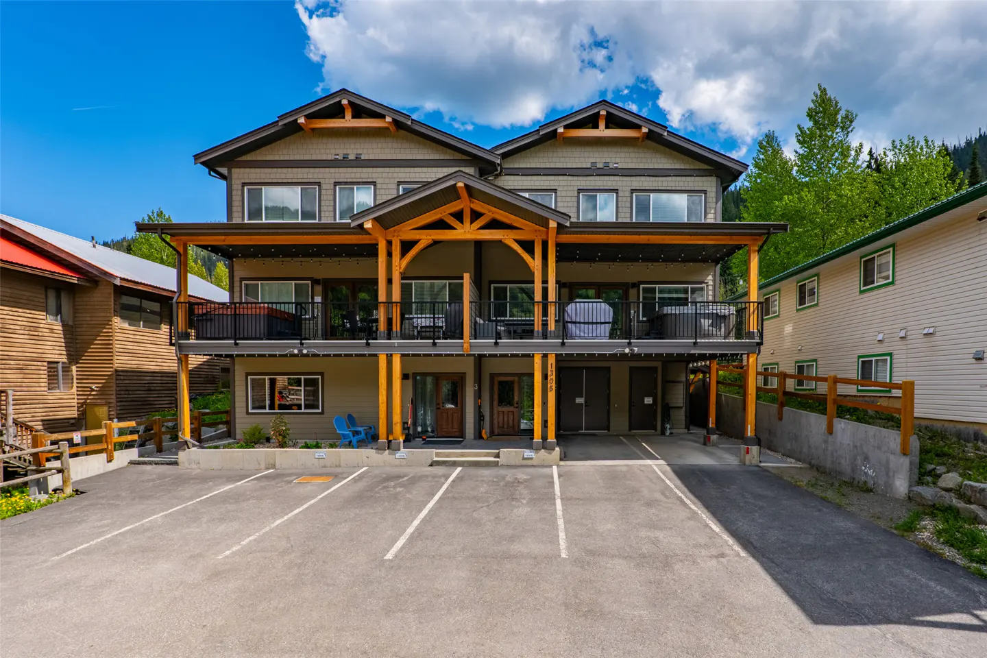 Three-story mountain lodge with a large wooden deck, hot tubs, and a paved parking lot under a bright blue sky.