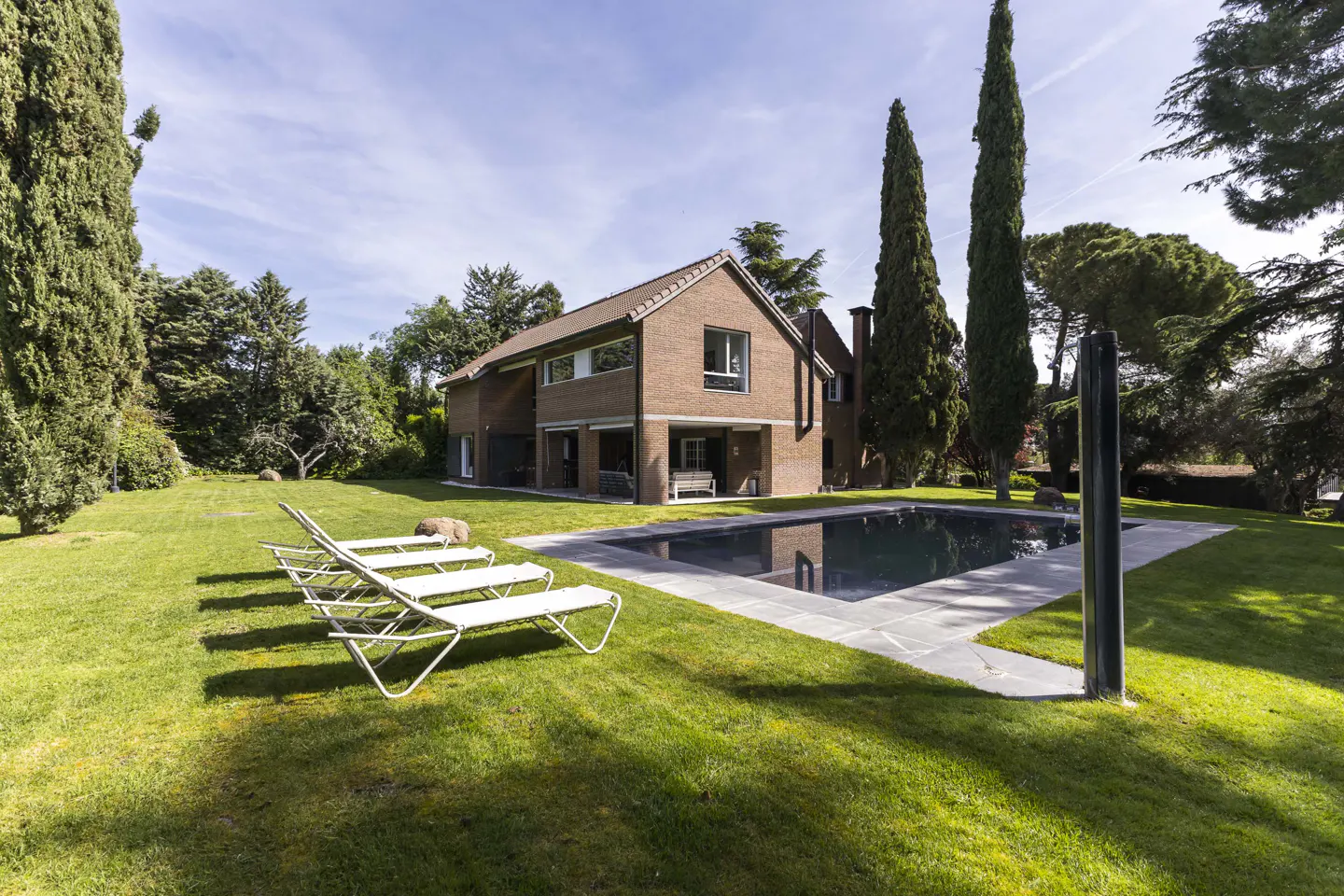 Brick house with a pool and lawn. Four white lounge chairs sit on the grass. Tall trees surround the property.