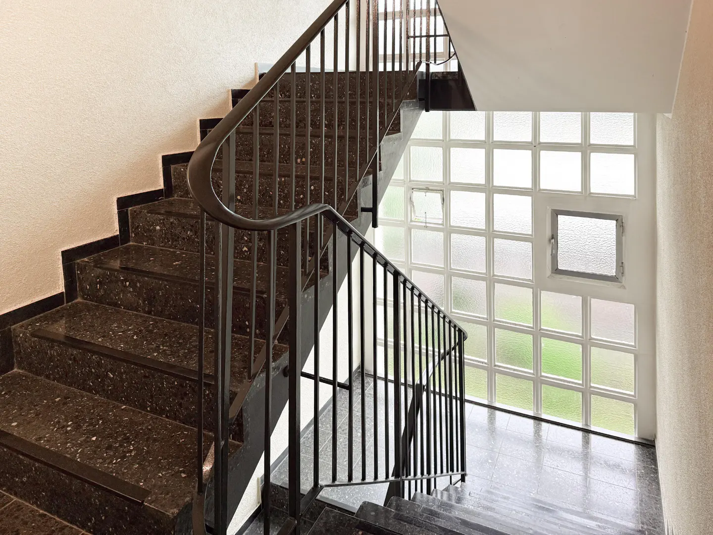 A staircase with black metal railings and speckled stone steps leads up to a wall of glass blocks.