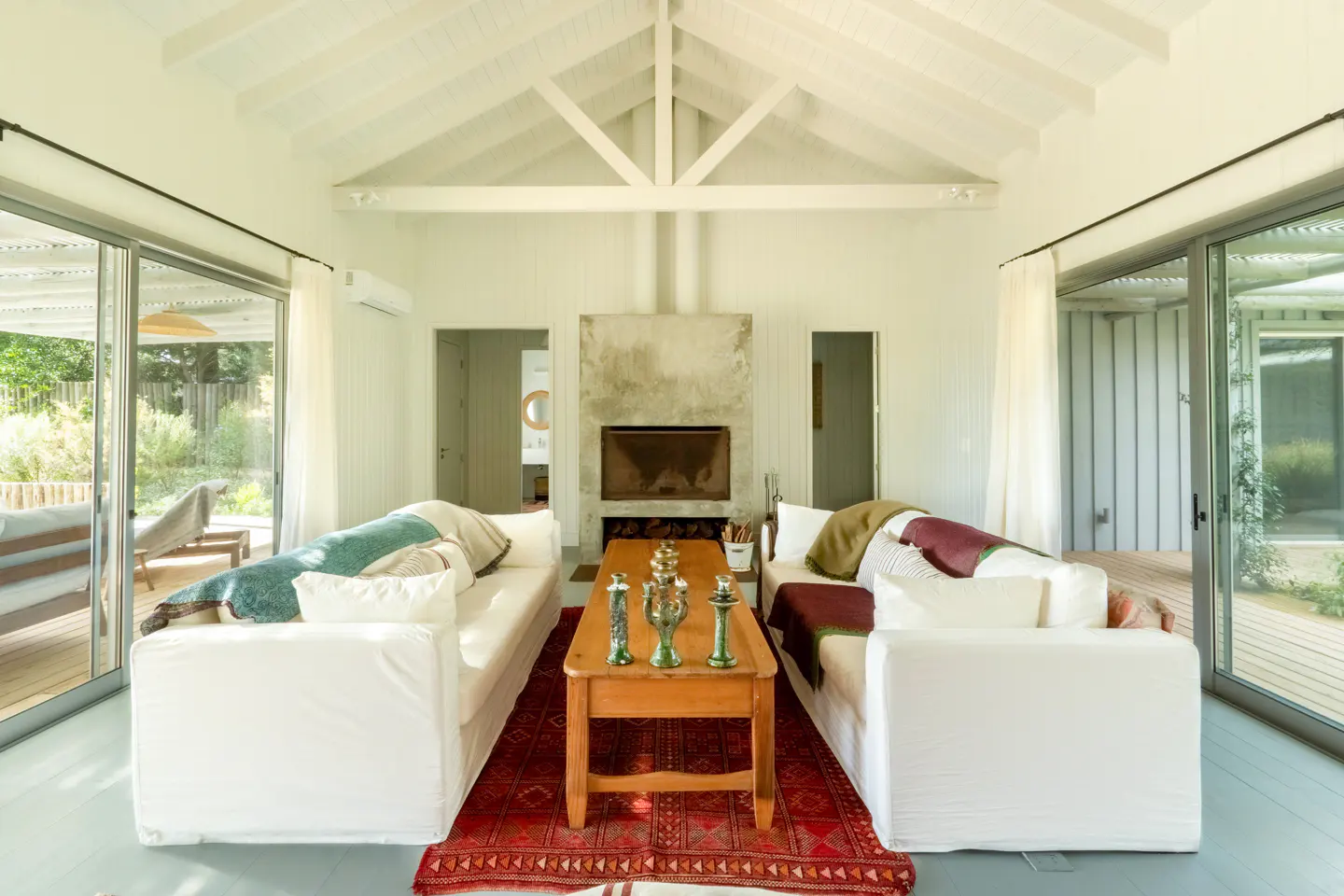 Bright living room with white sofas, a red rug, and a wooden coffee table with decorative objects. A fireplace is centered between doorways. Sliding glass doors lead to a deck.