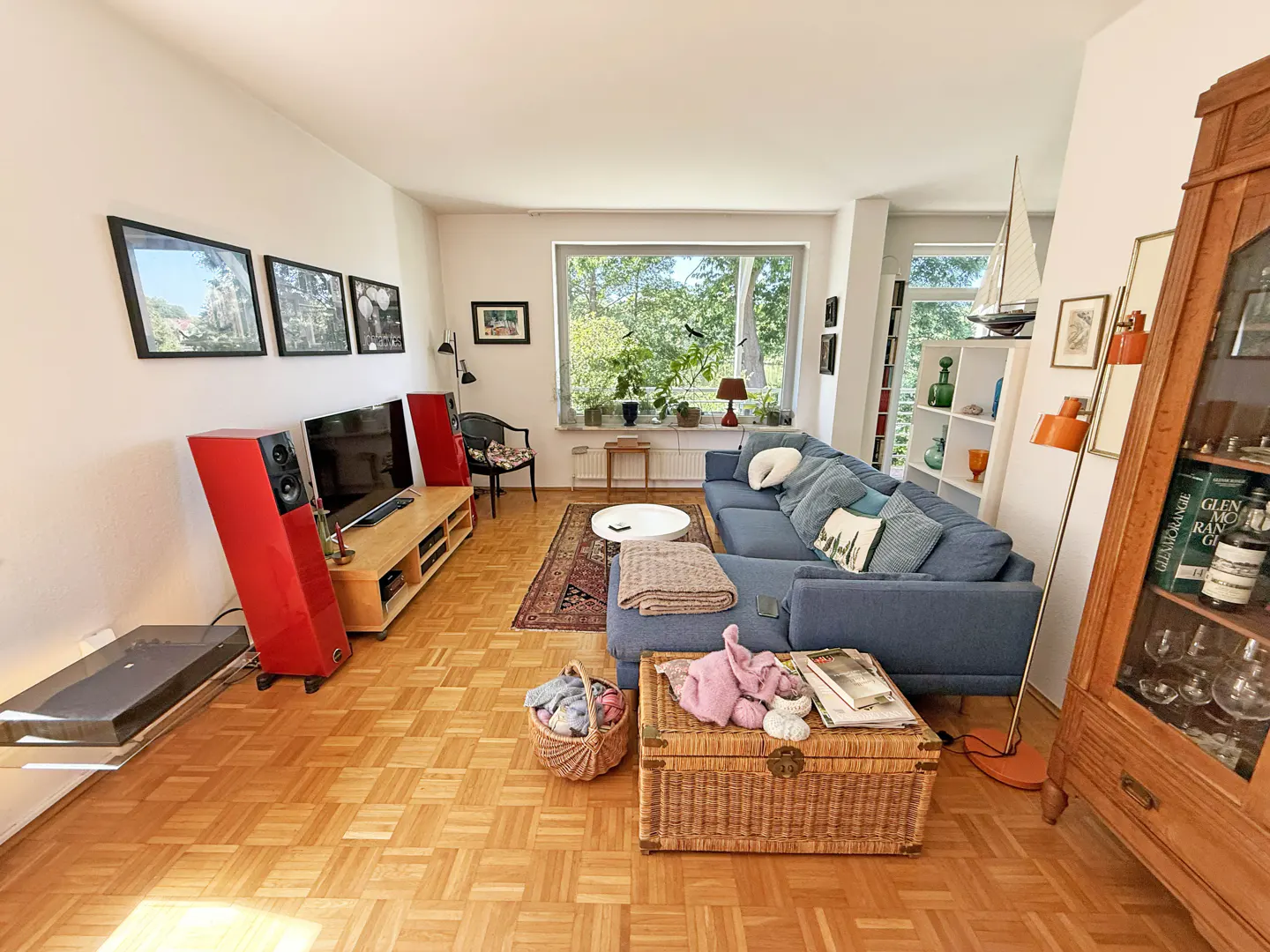 Bright living room with parquet floors, blue sectional sofa, red speakers, and a large window overlooking greenery.