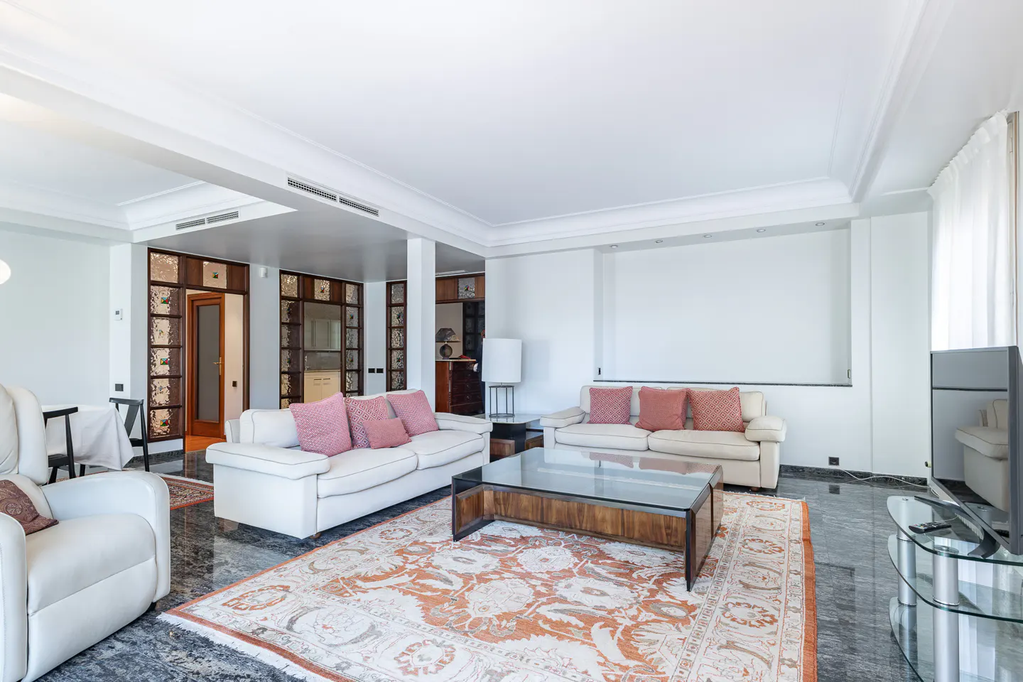 Bright living room with white sofas, pink pillows, and a glass-topped coffee table on an ornate rug. Marble floors and white walls.