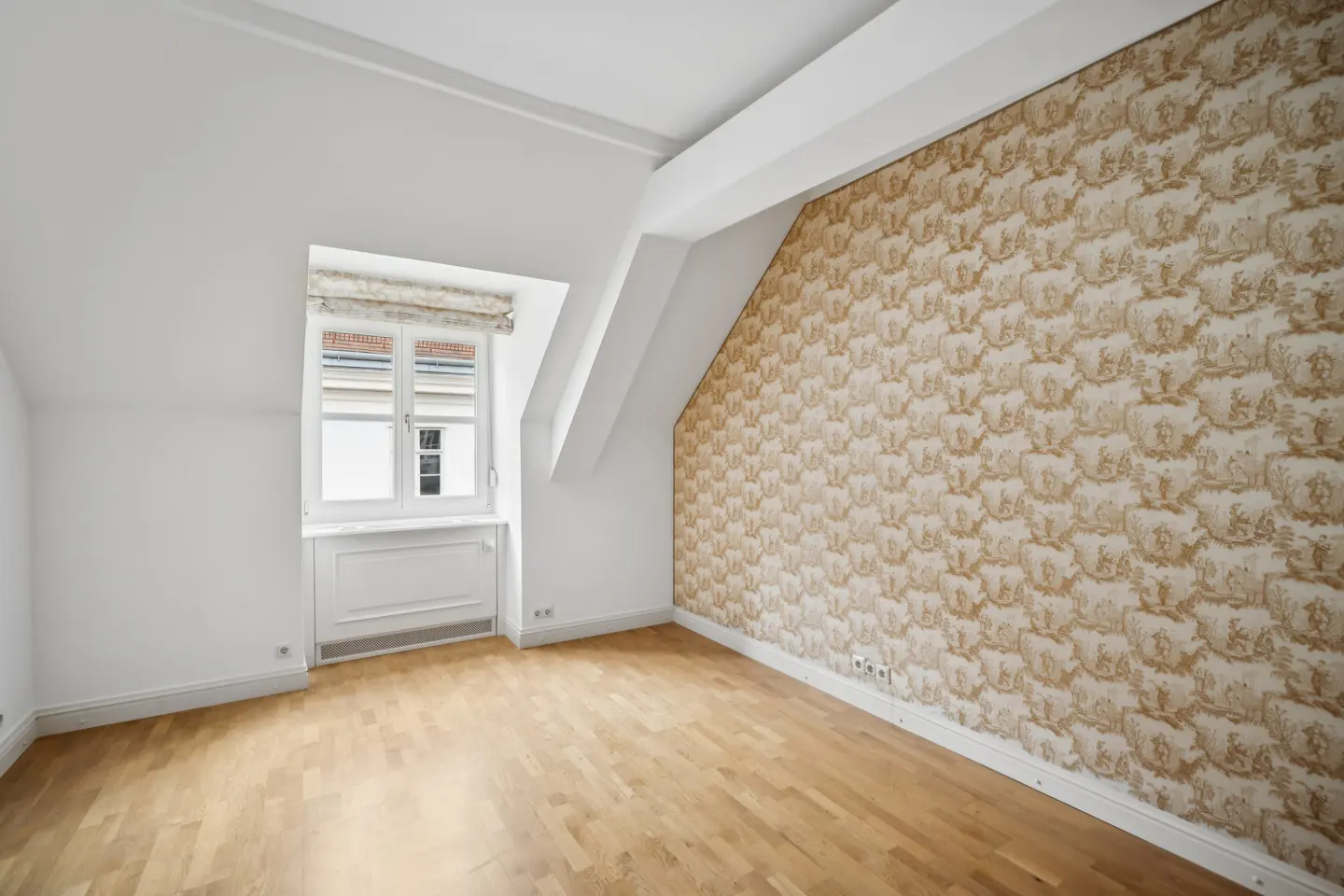 An empty room with light wood floors, white walls, and a patterned wallpaper accent wall. A window with a patterned shade is on the left.