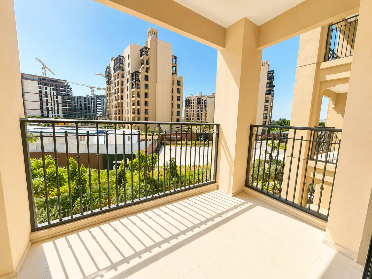 View from a beige balcony with black railings, overlooking buildings and green trees under a blue sky.