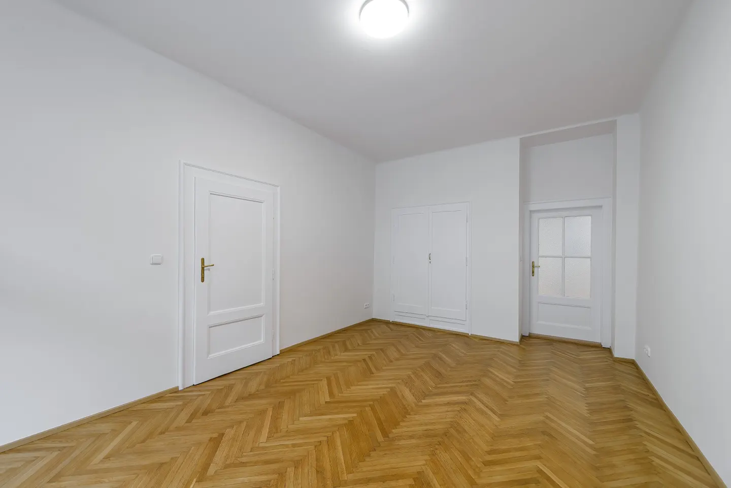 Bright, empty room with white walls, herringbone wood floor, and two white doors. A built-in closet is on the back wall.