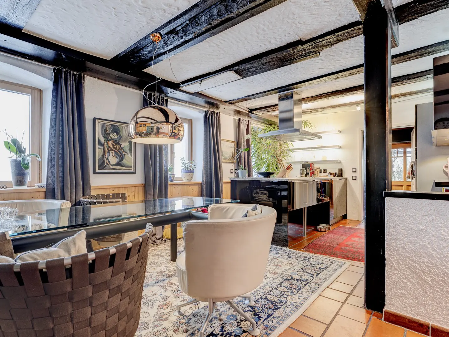 Open-concept kitchen and dining area with black beams, glass table, and modern chairs. A silver pendant light hangs above the table.