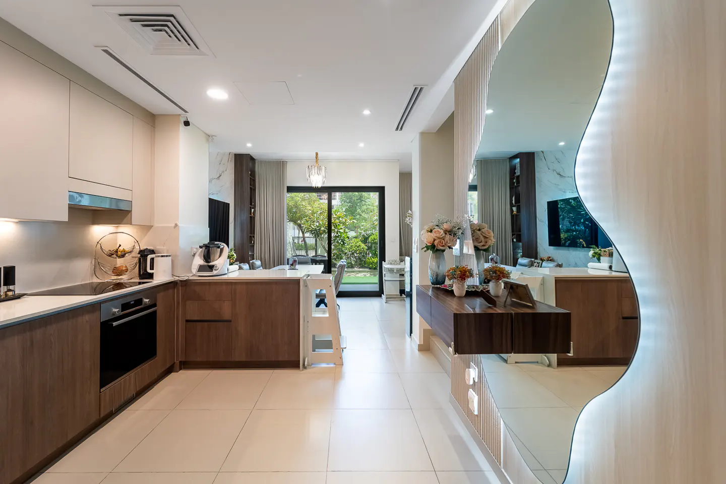 A modern kitchen with wood and white cabinets, stainless steel appliances, and a large mirror reflecting the living area. A child's step stool is near the island.