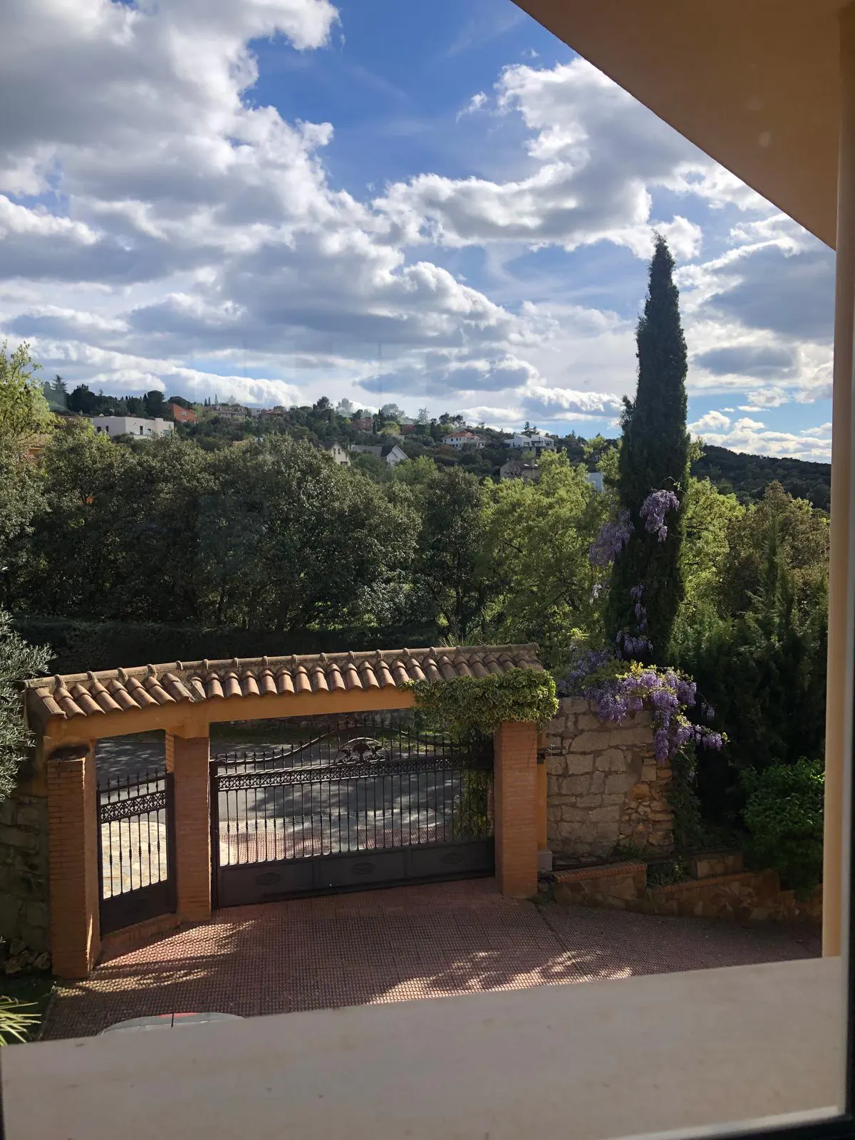 View from window of gated entrance to property. Brick pillars, tile roof, and black metal gate. Lush green trees and blue sky with clouds in background.