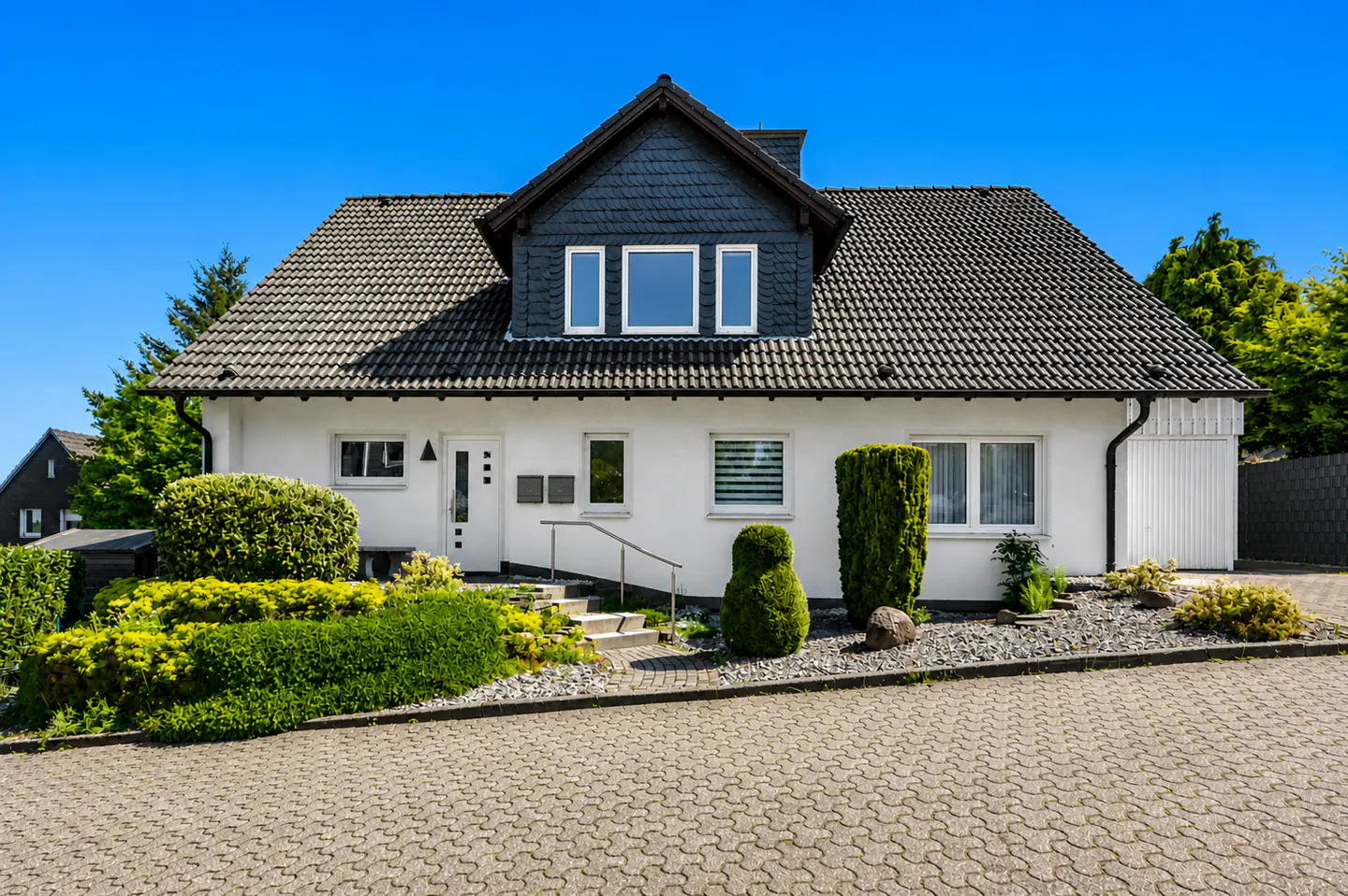 A white, one-story house with a gray roof and dormer windows under a clear blue sky. Green bushes line the front.