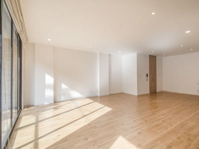 Bright, empty room with light wood floors, white walls, and large windows with tan blinds. Sunlight streams in.