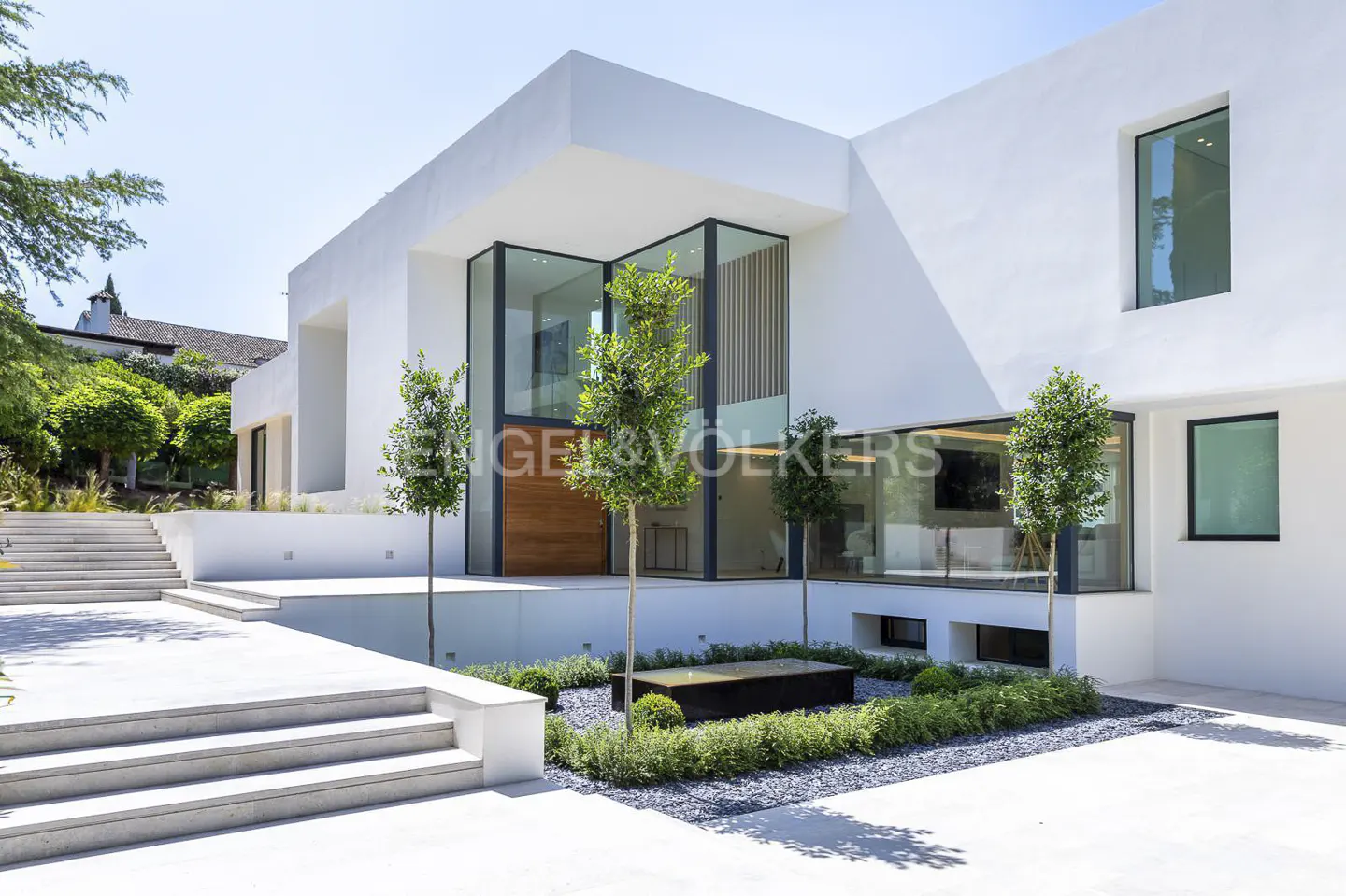 Modern white villa with large glass windows, a wooden front door, stone steps, and a landscaped courtyard with trees.