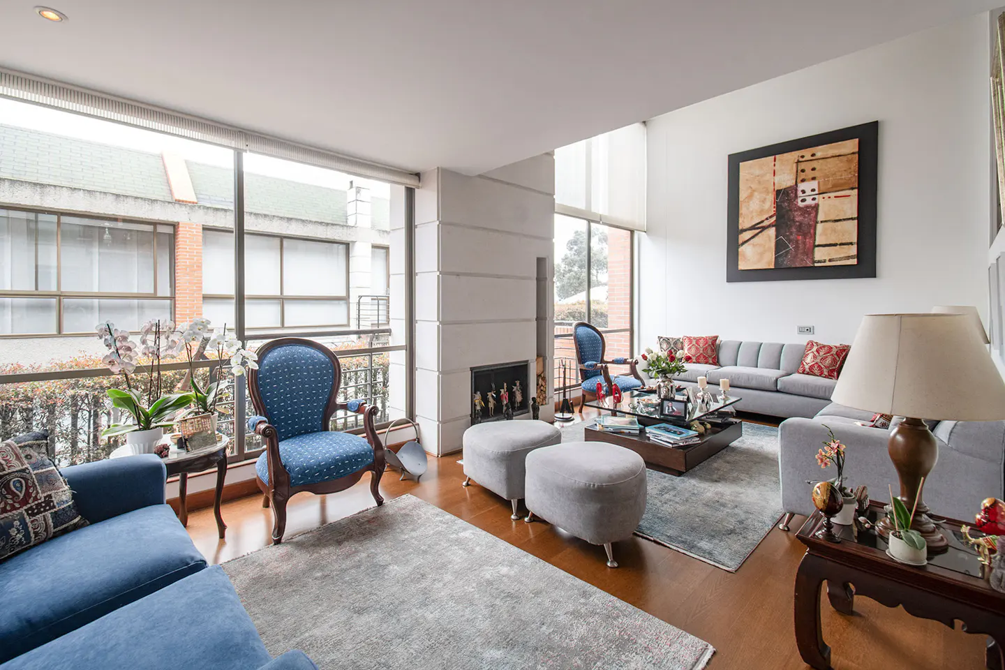 Bright living room with hardwood floors, blue sofas, gray rug, and large windows overlooking a balcony. Modern art hangs on the white wall.