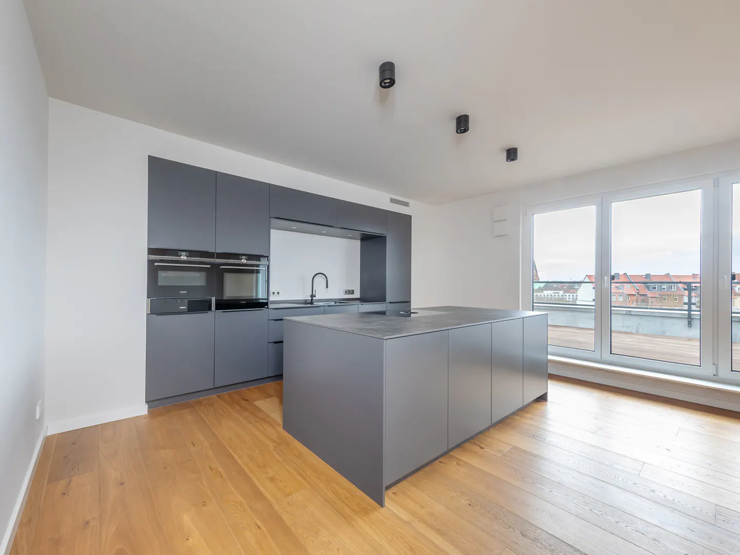Modern kitchen with gray cabinets, island, and wood floors. Large windows offer a view of rooftops.