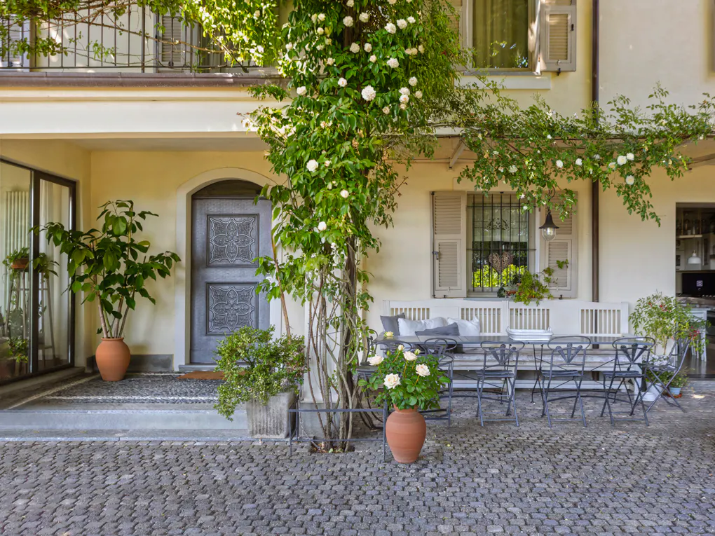Exterior of a cream-colored house with a gray door, stone patio, and climbing white roses. Outdoor dining set with a bench and chairs.