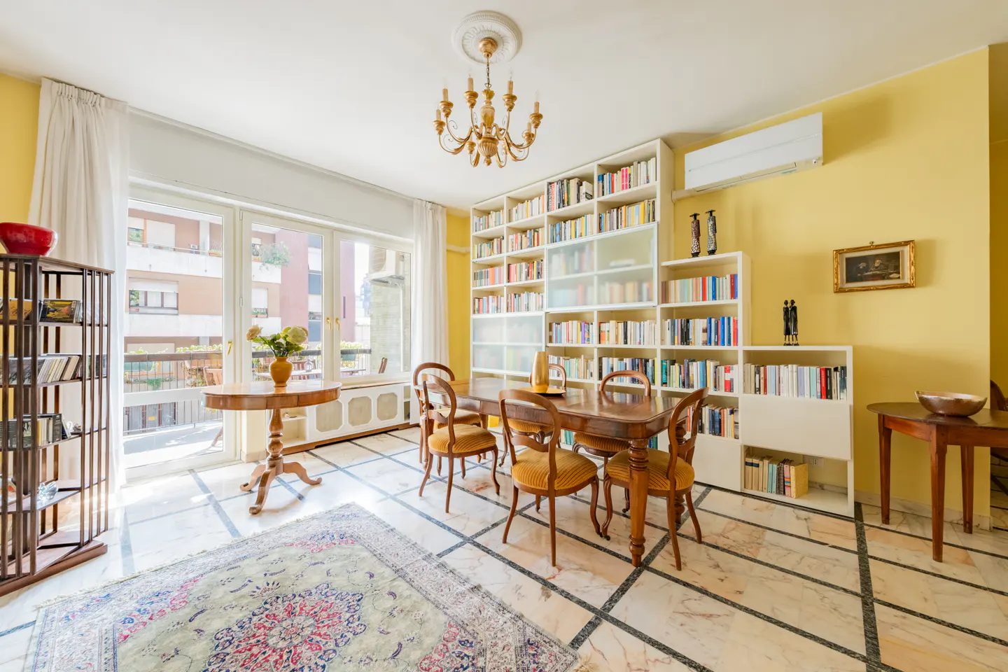 Bright living room with yellow walls, marble floors, and a large bookcase filled with books. A wooden table and chairs sit in the center of the room.