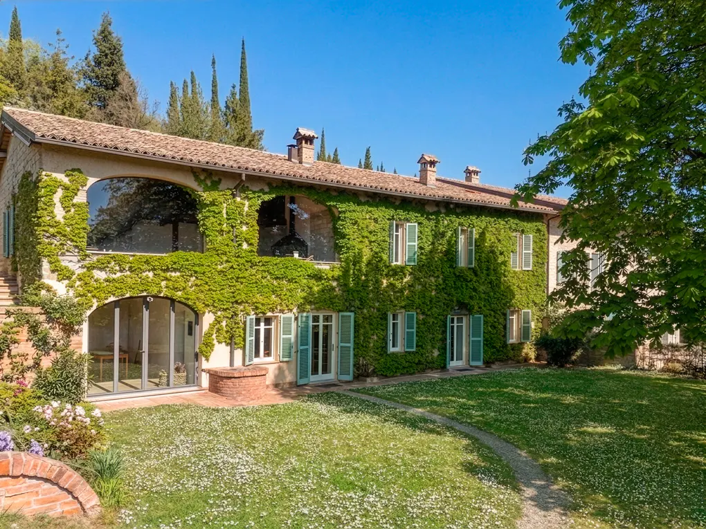Exterior view of a two-story house covered in green ivy, with light green shutters and a tiled roof, surrounded by a green lawn.